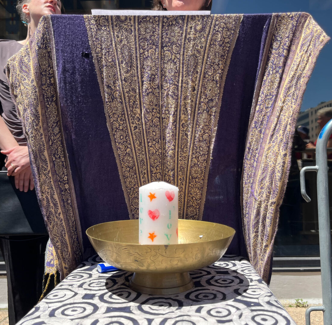 A thick white candle with the word “love” stamped onto it in green ink twice, three red hearts painted onto it, and two orange stars painted onto it sits in the center of a wide golden chalice with abstract engravings on its sides. The chalice is on a table covered in a dark indigo tablecloth with thick, white three-ringed ripple shapes printed on it. A blue plastic lighter is partially tucked under the chalice. A dark purple shawl with golden paisley patterns running down the middle and sides is draped up behind the chalice. There is a small hole towards the top of the shawl, and it has small, braided golden and purple tassels at the bottom. This forms an altar. The chin, brown hair, and lower ears of a white person with a small jade earring can be seen as they read from a white piece of paper. To the right of the person reading, a large glass window reflects a blue sky and two people, one of them with gray hair and another wearing a navy blue bucket hat, can be seen looking towards the person reading. The edge of a steel crowd control fence with a blue spraypaint mark is visible on the righthand edge of the image. To the left of the person reading, a white woman with short hair has her chin tilted up and eyes closed. She is wearing metal hoop earrings with a splash of teal in the center, a dark gray T-shirt, a silver wedding band, and black pants. She holds a black leather folder in her crossed hands.
