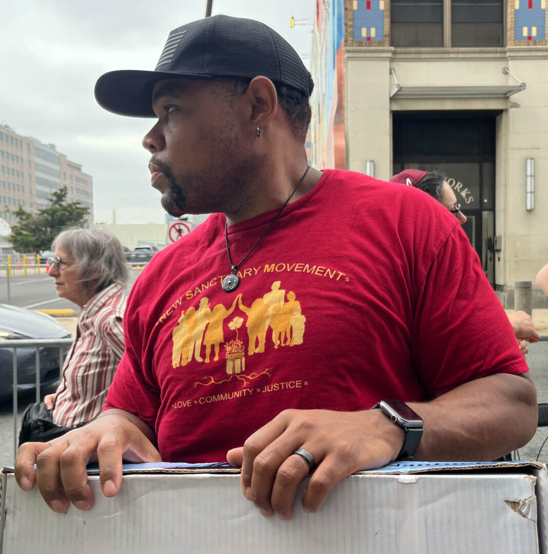 A lefthand side profile portrait of a middle-aged Black man with a goatee and lightly buzzed hair wearing a black baseball cap with seven lines on the front, a silver earring, a silver compass pendant, a red New Sanctuary Movement of Philadelphia T-shirt, a black smartwatch on his left wrist, and a black wedding band. The T-shirt has yellow and pale orange ink, reads “NEW SANCTUARY MOVEMENT” across the top and “LOVE, COMMUNITY, JUSTICE” across the bottom. Between the wording, there is a graphic that shows the outlines of eight people, some of whom appear to be children high-fiving, as they stand around a potted hibiscus flower with vegetation coming out of the top of the pot and roots coming out of the bottom. The man is holding a flat white cardboard box as he looks to his left. Behind the man and on his left, a white person with a gray bob haircut and circular glasses wears a red and cream striped button-up shirt and a black fanny pack. They are facing the same way as the man. Over the man’s right shoulder, a white woman’s nose, eyeglasses, forehead, black and white hair, and red yarmulke are visible. She is lifting her hands slightly. The sky in the background is light gray and overcast. In the lower lefthand corner of the image, a black car passes by behind the people.