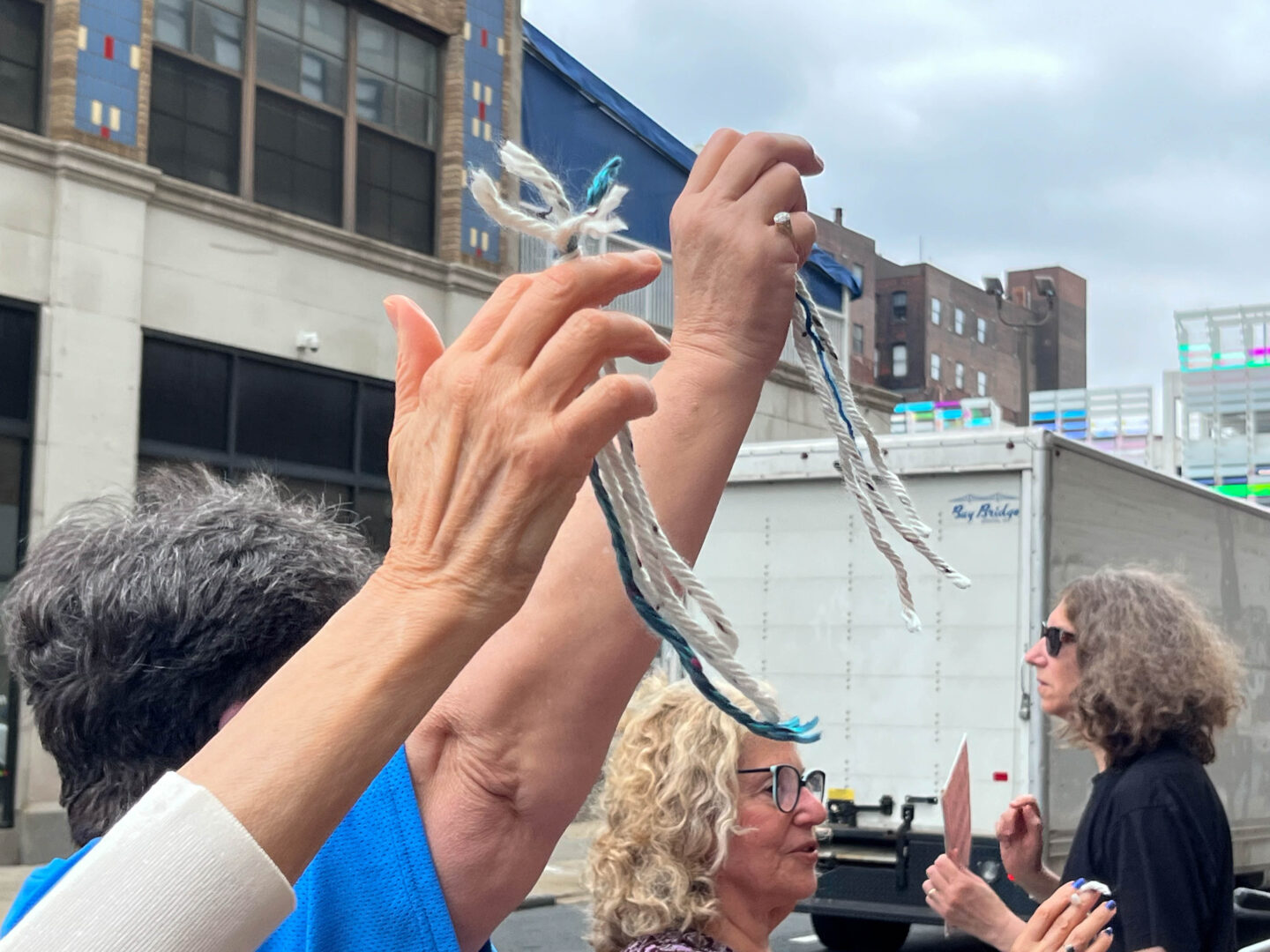 Two light-skinned hands each raise a tied knot of three white pieces of yarn and one blue piece of yarn. This knot is called a tzitzit. One of the hands has a cream-colored cardigan cuff, and the other one wears a diamond pinky ring. The person with the ring is wearing a blue jersey T-shirt and has a gray pixie cut. To the right of the raised hands, an older white woman with curly blond hair, blue-rimmed glasses, and blue nail polish also raises tzitzit in front of her as she speaks, and another white woman, who has graying light brown hair and is wearing sunglasses and a black T-shirt, holds a paper sign so that it faces the street behind the people with the tzitzit, where a white delivery truck drives past. The building across the street has floor-to-ceiling windows on the ground floor, and is made of tan brick and stone, with some colored tiles. It has a blue tarp draped over a fenced second-level area, and further down the street, a parking garage with colorful glass panels and a taller brick building can be seen in the distance.