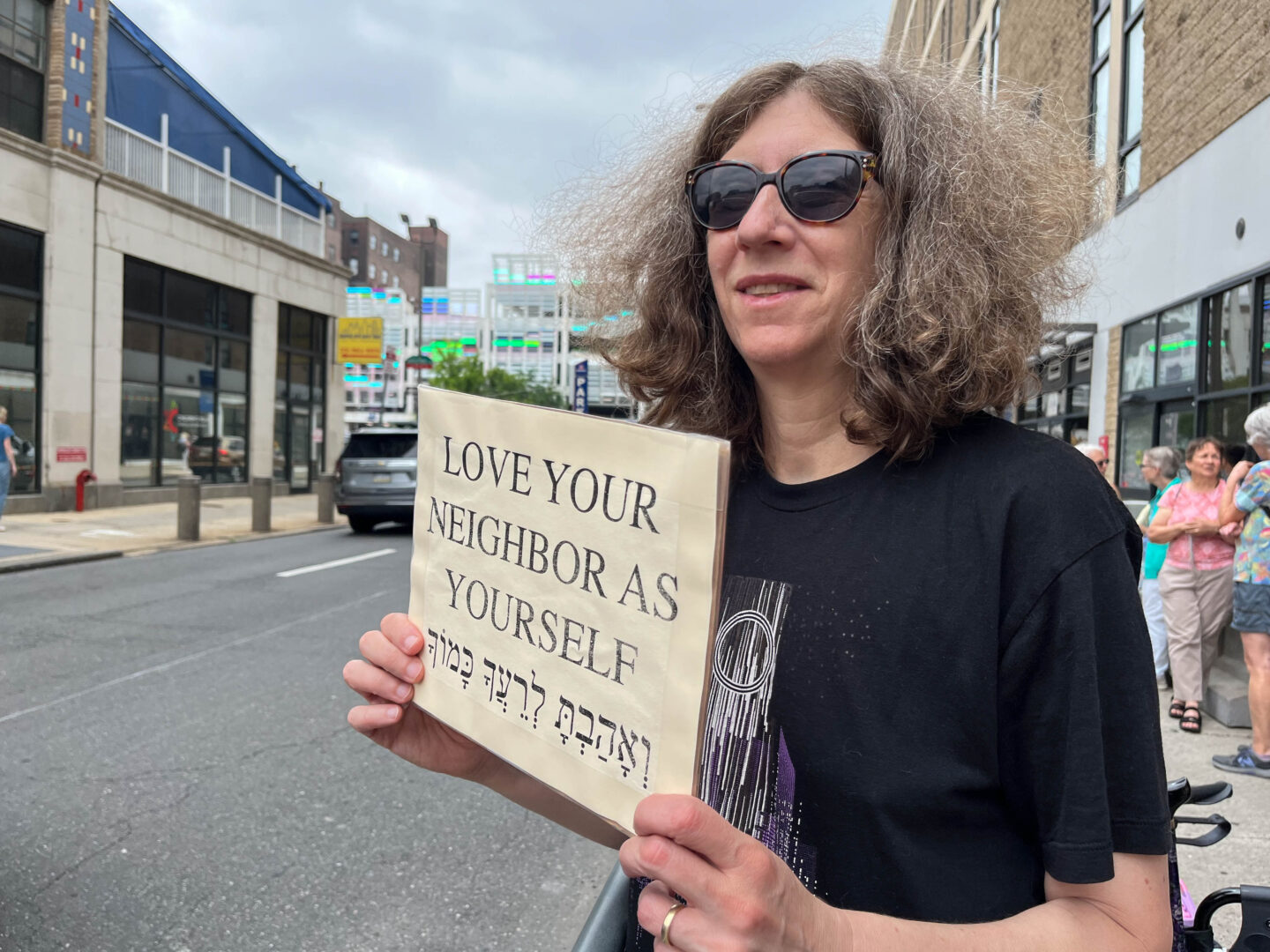A middle-aged white woman with curly, graying light brown hair smiles as she holds a sign that reads “LOVE YOUR NEIGHBOR AS YOURSELF” in both English and Hebrew. She is wearing wide sunglasses with brindle rims, a black T-shirt with an abstract white and purple pattern on it, and a golden wedding band. She is standing between a steel crowd control fence on her right and a parked walker on her left. Four white women in colorful shirts, three with gray or white hair, are talking a few feet behind the woman with the sign. The sign is pointed towards the street, which is to the right of the woman holding it. There are tan stone buildings with ground-level floor-to-ceiling glass windows on both sides of the street, and the multicolored glass exterior of a parking garage shines further down, with a taller red brick building behind it in the distance. The building across the street has a blue tarp draped over a fenced second-level area. A gray mini van is parked across the street from the woman holding the sign, and the top of a tree peeks out above the sign.